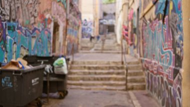 Grafiti kaplı duvarlar ve varoş mahallesindeki çöp kutularıyla dolu bulanık şehir sokakları, odak noktasından uzak figürlerle renkli ve kaotik bir atmosfer yaratıyor..