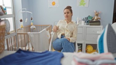 Bebek odasında beşik oyuncaklarıyla oturan kadın, günlük kıyafetlerle rahatlıyor, rahatlatıcı bir dekorla çevrili, bir sükunet hissi uyandırıyor, pencereden içeri güneş ışığı giriyor..