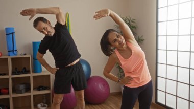 Orta yaşlı İspanyol çift kapalı spor salonu ortamında birlikte esneme hareketleri yapıyor, fitness, egzersiz, sağlık ve ortaklığın altını çiziyor.