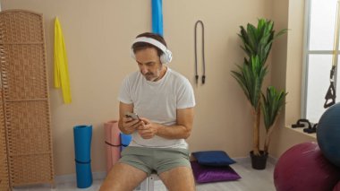 SağIık odasında akıllı telefon kullanan, spor aletleriyle çevrili, rahat ve odaklanmış görünen, kulaklık takan, yoga minderleri ve bitkilerle evde oturan bir adam..