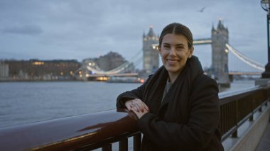 Londra 'da köprüde gülümseyen genç İspanyol kadın. Arka planda bulutlu bir gökyüzünün altında köprü var. Birleşik Krallık' ta bir kış manzarası sergiliyor..