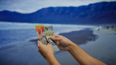 Manzaralı bir plajda Avustralya banknotlarını tutan eller, deniz kenarı zeminiyle finansal bir konsepti tasvir eden, para birimi ve yaz rahatlama unsurlarını birleştiren eller..