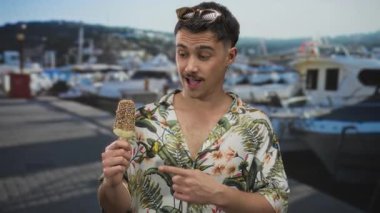 Genç adam arka planda teknelerle deniz kenarındaki bir limanda dondurmanın keyfini çıkarıyor. Açık hava tişörtünün içinde sıradan bir turist havası yaratıyor..