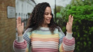 Kıvırcık saçlı İspanyol kadın, elleri renkli bir süveterin üzerinde, büyük jestler yapıyor, açık havada duruyor, neşe ve kendine güven gösteriyor..