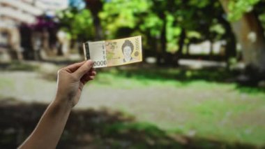 El ele tutuşan Güney Koreli, canlı yeşillik sergilenen güneşli bir parkta banknot kazandı. Bu da açık havada finansal bir tema olduğunu gösteriyor..