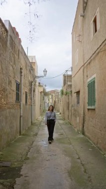 Bir kadın, Mallorca 'nın manzaralı eski bir şehir sokağında mutlu bir şekilde geziniyor, kırsal Akdeniz mimarisi ve enerjik enerjisiyle çevrili, bu tarihi çevrenin cazibesini kucaklıyor..