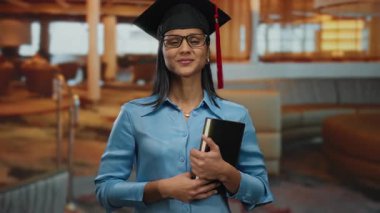 Mezuniyet şapkalı İspanyol kadın modern otel lobisinde elinde kitap, kameraya güvenle gülümsüyor, ev içi profesyonel ortamdaki başarıyı ve başarıyı simgeliyor..