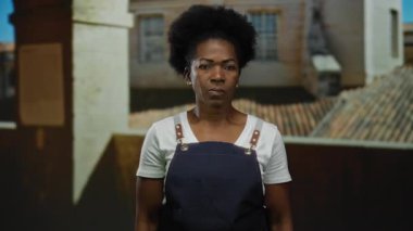 Eski bir üniversitede kapalı alanda önlük giyen bir kadın tarihi bir mimariye karşı ciddi bir ifadeyle bir jest yapıyor..