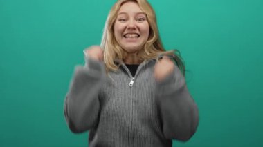 Yeşil arka planda sevinçle kutlama yapan bir kadın gri süveter giyiyor. Mutluluk heyecanı ifade ediyor. Enerjik bir hava.