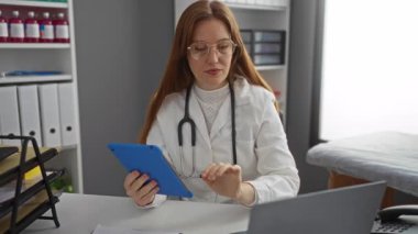 Klinikte tablet tabletle çalışan bir kadın. Tıp alanında çalışıyor. Modern çalışma ortamında kızıl saçlı çekici bir doktor, dosyalar ve ekipmanlarla çevrili..