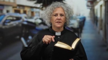 Gri saçlı yaşlı bir kadın bir şehir caddesinde kitap tutuyor ve kutsal bir şehir anını ruhani bir odak ile yakalıyor..