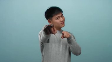 Genç adam güvenle gülümsüyor ve özgüvenli bir mavi arka plana işaret ediyor, bir stüdyo ortamında neşeli ve çekici bir tavır yakalıyor..