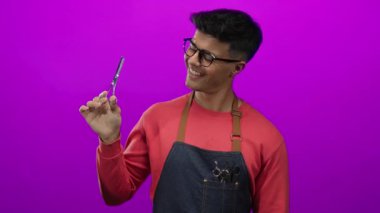 Genç, İspanyol asıllı bir adam parlak pembe bir arka plana karşı kuaför makası tutuyor. Gözlüklü, kırmızı kazaklı ve kot önlüklü, modern berber tarzı giyiniyor..