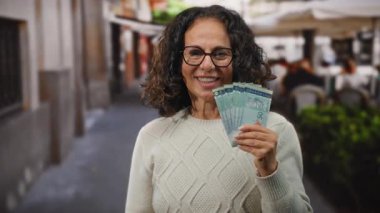 Orta yaşlı bir kadın bir restoranın terasında elinde Malezya sirkesi banknotları tutarken gülümsüyor, elinde para olan neşeli bir açık hava sahnesi sergiliyor..