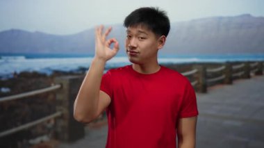 Kırmızı tişörtlü genç Çinli bir adam deniz kenarında sakin bir sahilde ve arka planda dağlarla hareket edebiliyor. Rahatlığı ve dışarıdaki sükuneti somutlaştırıyor..
