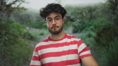 Çizgili tişörtlü genç sakallı bir adam açık havada, yemyeşil bir parkta mola veriyor..