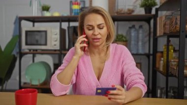 Pembe kazaklı kadın, ofiste mikrodalga fırın ve arka plandaki tabaklarla konuşurken kredi kartı tutuyor. Endişe ve odaklanma gösteriyor..