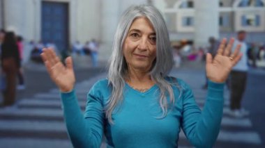 Gri saçlı yaşlı bir kadın hareketli bir açık sokakta duruyor, mavi giyinmiş ve hareketli bir şehir ortamında bulanık yayaların arasında kendini ifade ediyor..