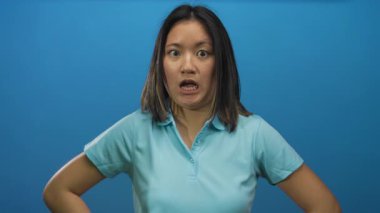 Mavi bir duvar arkaplanı üzerinde şaşkınlığını ifade eden, açık mavi bir gömlek giyen, çeşitli yüz ifadeleri sergileyen genç Çinli kadın şokta..