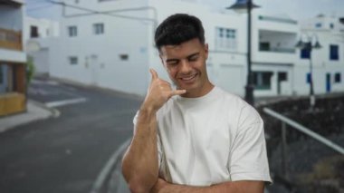 Şehir sokaklarında dikilen genç bir adam gülümseyerek bir telefon konuşması yapıyor, şehir ortamını ve günlük kıyafetleri vurguluyor..