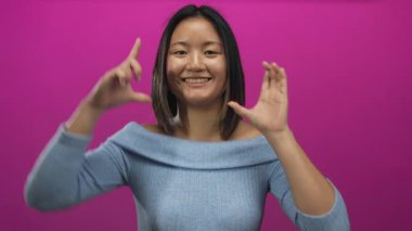 Genç Çinli kadın gülümsüyor ve elleri çerçeveli poz veriyor. Stüdyoda izole edilmiş pembe arka plana karşı..