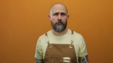 Turuncu arka planda kahverengi önlüklü sakallı bir adam ciddi bir ifade sergiliyor..