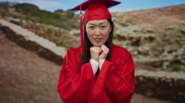 Kadın mezuniyeti kırmızı cübbe ve şapkayla kutluyor park ortamında neşeli bir ifadeyle.