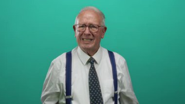 İş kılığındaki son sınıf öğrencisi yeşil arka plana yumruk atıyor, gözlük, kravat ve pantolon askısı takıyor, duygularını izole bir ortamda ifade ediyor..