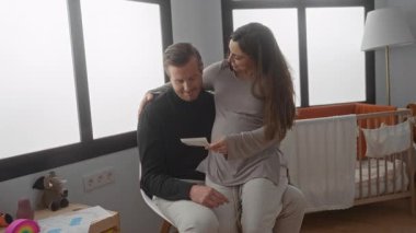 Hamile kadın ve erkek rahat bir odada oturup beşiğin yakınındaki ultrason fotoğraflarını inceliyor ve gelecekteki bebekleri için heyecanlı olduklarını gösteriyorlar..
