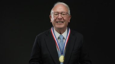 Madalya takmış kıdemli bir adam siyah bir arkaplana öpücük yolluyor. Kutlama ve tanınma ayinini izole bir ortamda yapıyor..