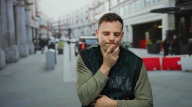 Canlı bir şehir caddesinde duran genç adam, arka planda bulanık şehir manzarasıyla düşünceli ve düşünceli görünüyor..