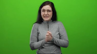 Parlak yeşil arkaplan önünde karışık duygular ifade eden kadın kumral saç, gözlük ve süveter sergiliyor..