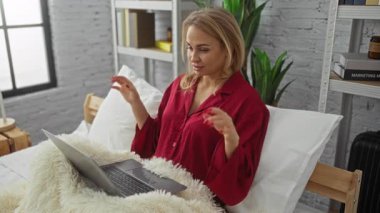 Yatak odasında dizüstü bilgisayar kullanan, kırmızı pijama giyen çağdaş dekorlu, modern ve rahat bir yaşam tarzı sergileyen bir kadın..