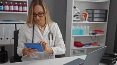 Hastane ofisinde tablet kullanan kadın doktor. Klinik ortamda çalışan tıbbi malzeme ve ekipmanlarla çevrili. Gözlük ve laboratuvar önlüğü takıyor..