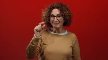 Kıvırcık saçlı, gözlüklü, kırmızı arka planda küçük boy, kendine güvenen, arkadaş canlısı bir ifade sergileyen bir kadın.