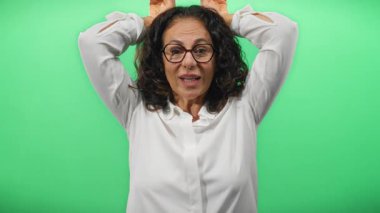 Yeşil duvarın önünde şakacı kulaklar yapan, gülümseyen bir kadın. İzole edilmiş arka plan. Beyaz bluz, kıvırcık saç gözlükleri, orta yaşlı İspanyol gözlüğü.