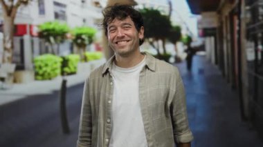 İspanyol asıllı bir adam açık havada yeşil ağaçlar ve kentsel arka planı ile canlı bir caddede dikilmiş, rahat ve yaklaşılabilir bir tavır sergiliyor..