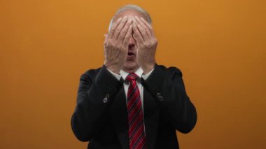 Takım elbiseli yaşlı adam yüzünü ellerle kapatıyor, sonra turuncu bir arka plan duvarına karşı neşeli bir gülümseme sergiliyor..