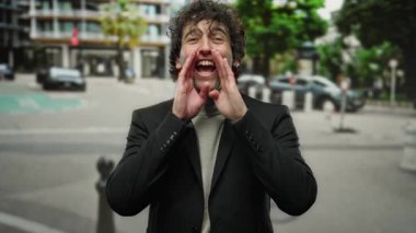 Şehir caddesinde siyah takım elbiseli bir adam bağırıyor, şehir dışında trafik ve binalarla çevrili bir ortamda duygularını ifade ediyor..