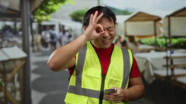 Güvenli yelek giyen bir adam, şehir kafeteryasının arka planında canlı şehir yaşamını gösteren masalar ve sandalyelerle akıllı telefon tutarken jest yapıyor..