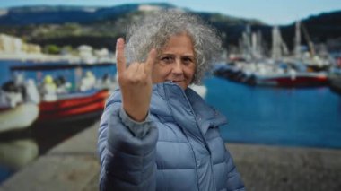 Orta yaşlı, gri saçlı bir kadın, deniz kıyısında bir kaya jesti yapıyor. Arka planda bulanık botlar ve dağlar olan mavi bir ceket giyiyor..