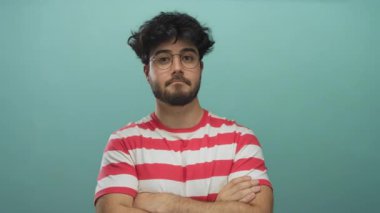 Mavi arka planda kollarını kavuşturan gözlüklü ve sakallı İspanyol genç adam çizgili gömlek giyiyor sakin bir ifade sergiliyor..