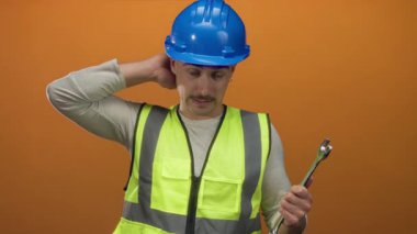 Yansıtıcı yelekli ve kasklı genç adam turuncu arka planda İngiliz anahtarı tutuyor, düşünceli görünüyor..