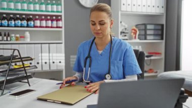 Klinikteki kadın steteskop ile önlük giyiyor. Ofiste notlar yazıyor. Etrafı raf ve ilaçlarla çevrili.