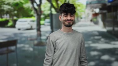 Sokak ortasında arka planda ağaçlarla gülümseyen, sıradan kıyafetler giyen İspanyol bir genç..