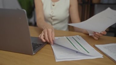 Modern bir ofis ortamında, dizüstü bilgisayar ve çizelgelerle mali belgeleri inceleyen, gözlük takan ve kolsuz bluz giyen bir kadın, temiz bir iç mekan ortamında çalışmaya odaklandı..