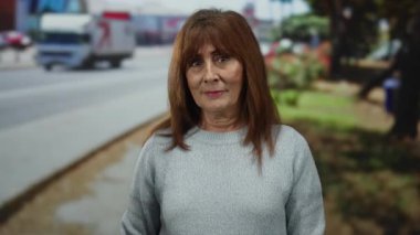 Latin kökenli son sınıf öğrencisi bir kadın şehir caddesinde, kazak giymiş, kafasına dokunarak jestler yapıyor, şehir trafiği ve arka planda ağaçlarla birlikte, tefekkür ya da farkındalık öneriyor..