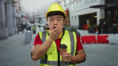 İnşaat işçisi, elinde sert şapka ve güvenlik yeleği olan bir kahveyle şehir sokaklarında esniyor..