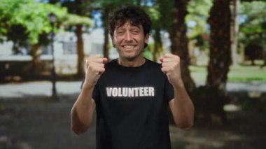 İspanyol bir adam yeşil bir parkta gönüllü tişört giyerek neşeli bir şekilde alkışlıyor. Güneşli bir ortamda cazibe ve mutluluk saçıyor..