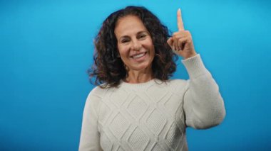 Kıvırcık saçlı İspanyol kadın, mavi arka plandaki el hareketlerini kullanarak duygularını ifade ediyor. Ciddi bir ifadeyle baş parmağından olumlu bir başparmağa geçerken gülümsüyor..
